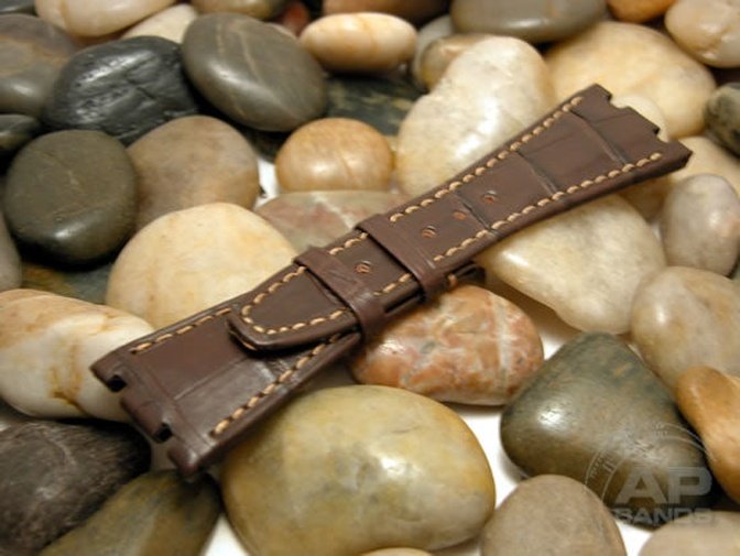 Brown crocodile leather watch strap with beige stitching on pebbles.
