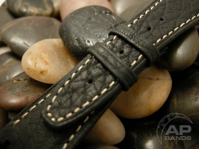 Black leather watch strap with white stitching rests on smooth stones.