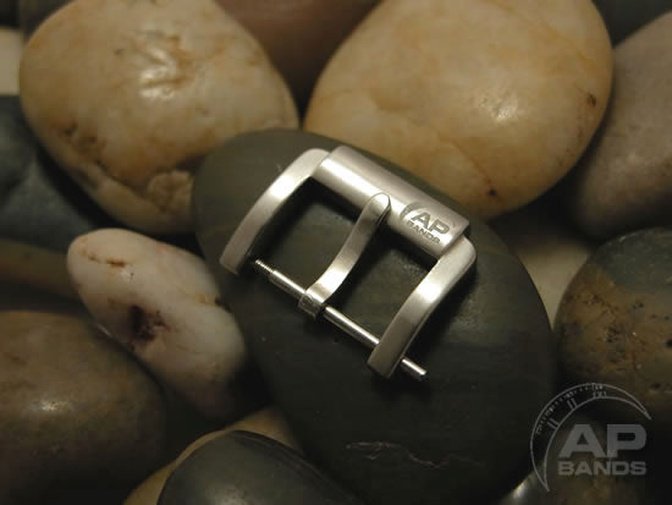 A brushed metal AP Bands watch buckle rests on a smooth, dark stone.