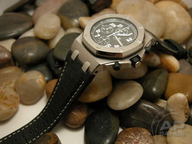 A stainless steel chronograph watch with a black leather band rests on a bed of smooth river rocks.