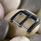 Gunmetal watch buckle with engraved AP Bands logo rests on smooth stones. 
