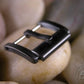 Black PVD watch buckle resting on smooth stones. 
