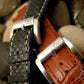 Black checkered carbon fiber and brown alligator leather watch straps with brushed metal buckles rest on smooth stones. 
